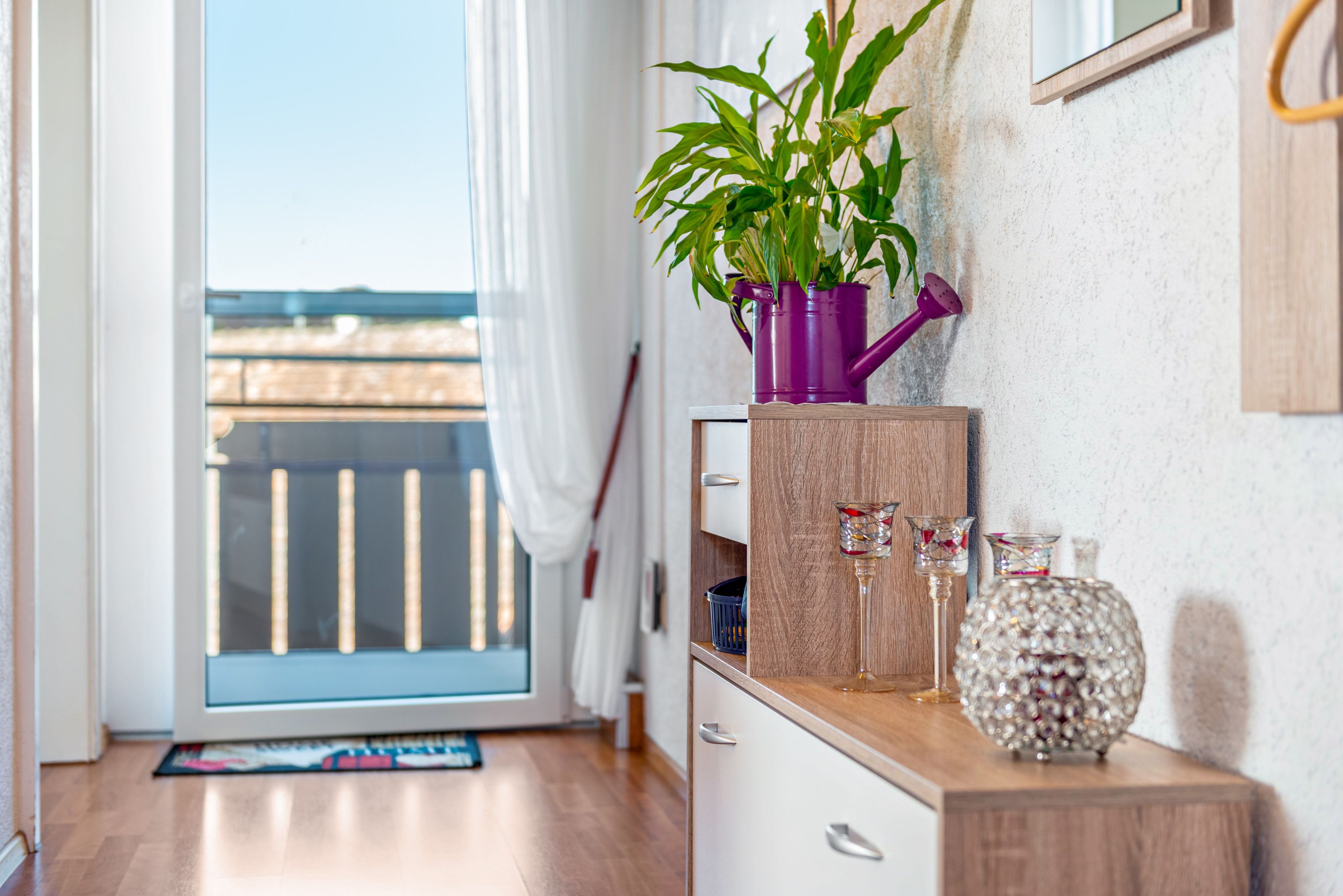 A room with a wooden cabinet, a purple watering can, a potted plant, and decorative glass items.