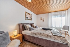 Un dormitorio con techo de madera, una cama con muchos cojines, una mesita de noche con una lámpara y una silla.