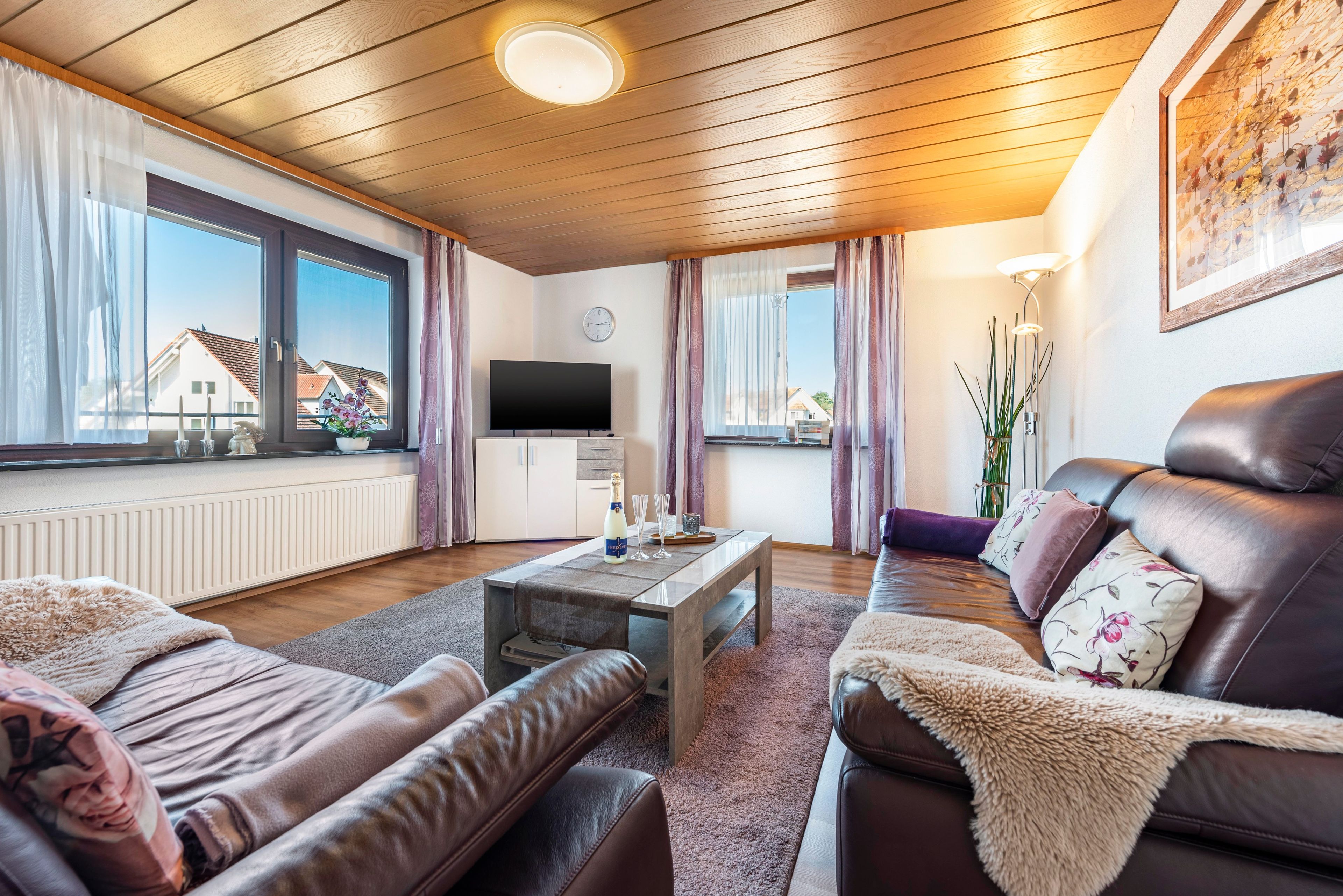 Ein modernes Wohnzimmer mit einer Couch, einem Couchtisch und einem Fernseher. Es gibt ein Fenster mit Vorhängen, eine Heizung und eine Uhr an der Wand.
