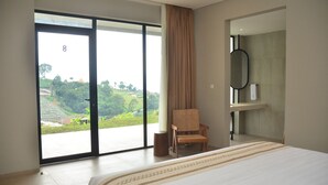 Camera da letto con un letto grande, una sedia, bagno con specchio e vista su un paesaggio verde attraverso una porta scorrevole.