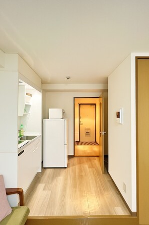 A compact kitchen with a refrigerator, a sink, and a microwave. A wooden floor and a door leading to a hallway are also visible. - Casa G.S. Ebisucho (Osaka)