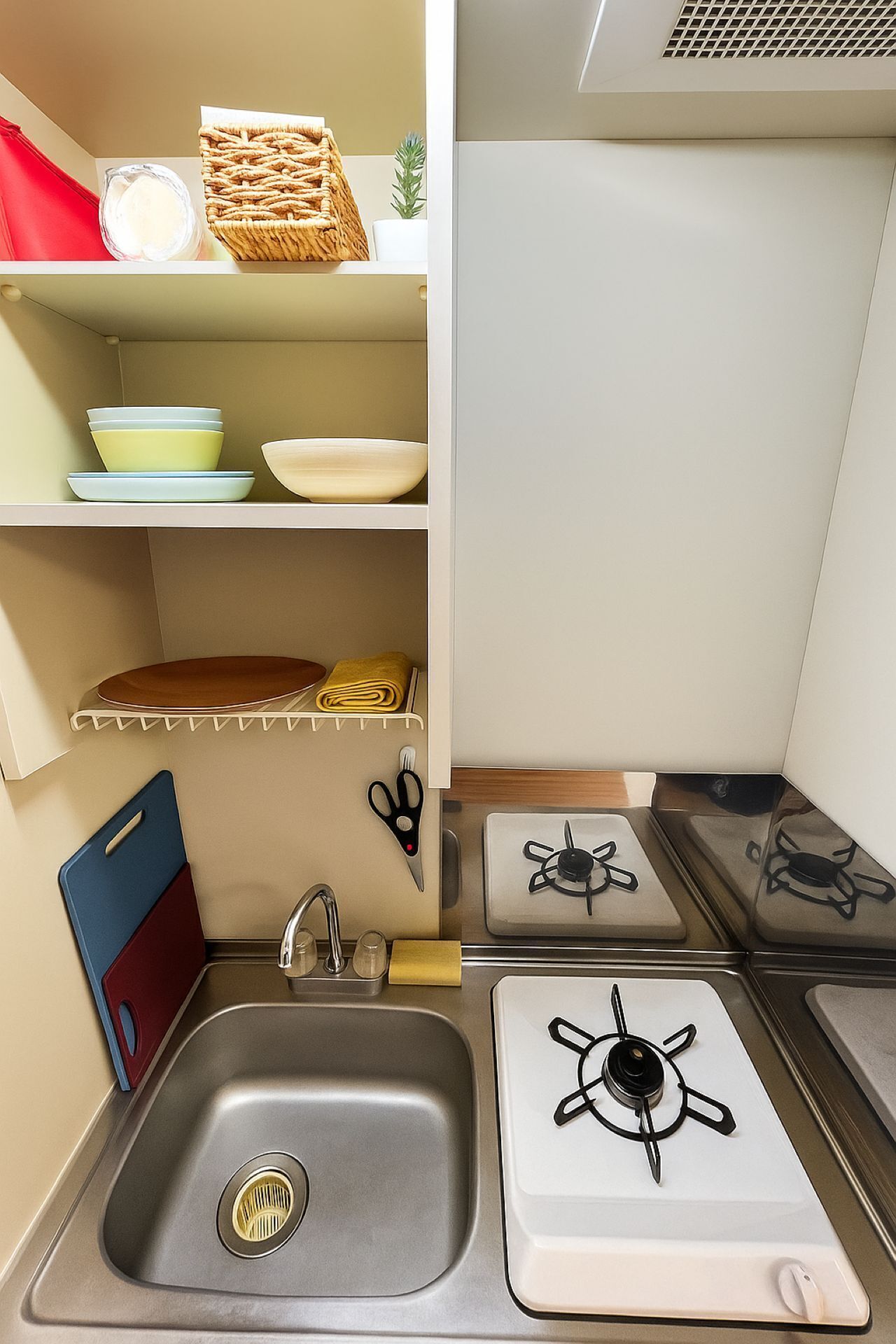 A compact kitchen with a sink, two stovetops, and shelves with dishes and kitchen utensils.