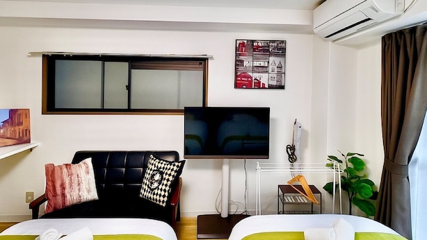 Ein Hotelzimmer mit zwei Betten, einem Fernseher, einer Klimaanlage, einem Fenster mit Jalousien, einem kleinen Schreibtisch und einer Pflanze.