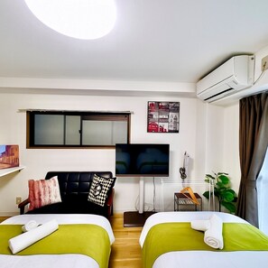 A hotel room with two beds, a TV, an air conditioner, a window with blinds, a small desk, and a plant. - Casa G.S. Ebisucho (Osaka)