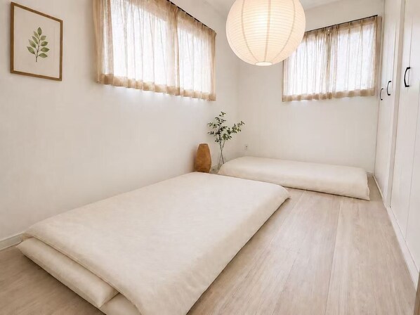 Quarto minimalista com duas camas, cortinas leves, luminária pendente e uma impressão botânica emoldurada.