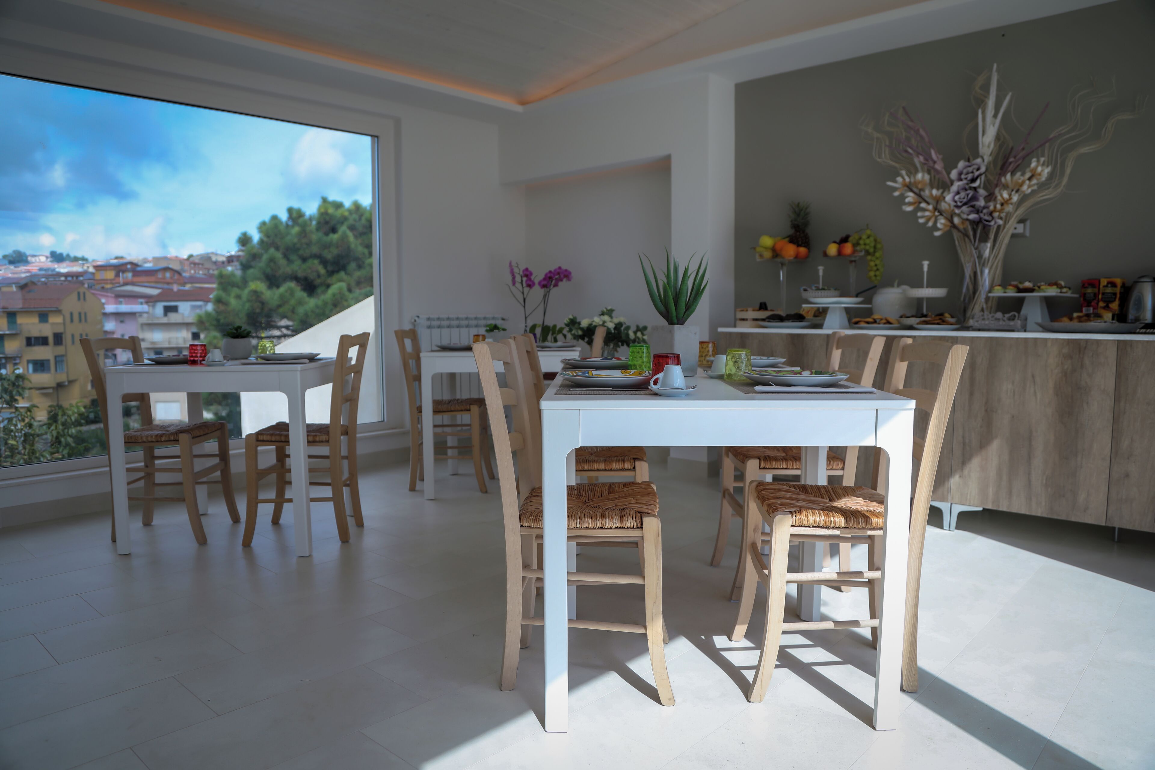 Un comedor moderno con una mesa blanca y sillas de madera, un ventanal con vistas a la ciudad y una encimera de cocina con diversos objetos.