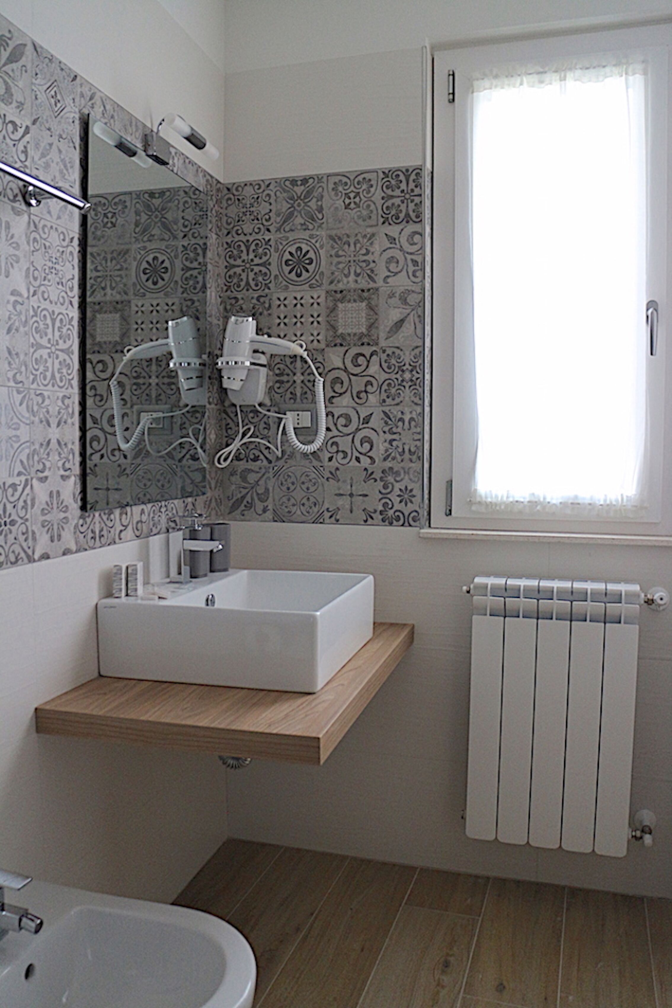 A bathroom with a white sink, a mirror, a towel rack, a shower head, and a window with a white curtain.