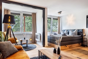 A modern bedroom with a bed, a sofa, a coffee table, a lamp, and a view of trees through large windows.