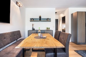 A modern dining area with a wooden table, dark upholstered chairs, a mounted flat-screen TV, and a decorative shelf above.