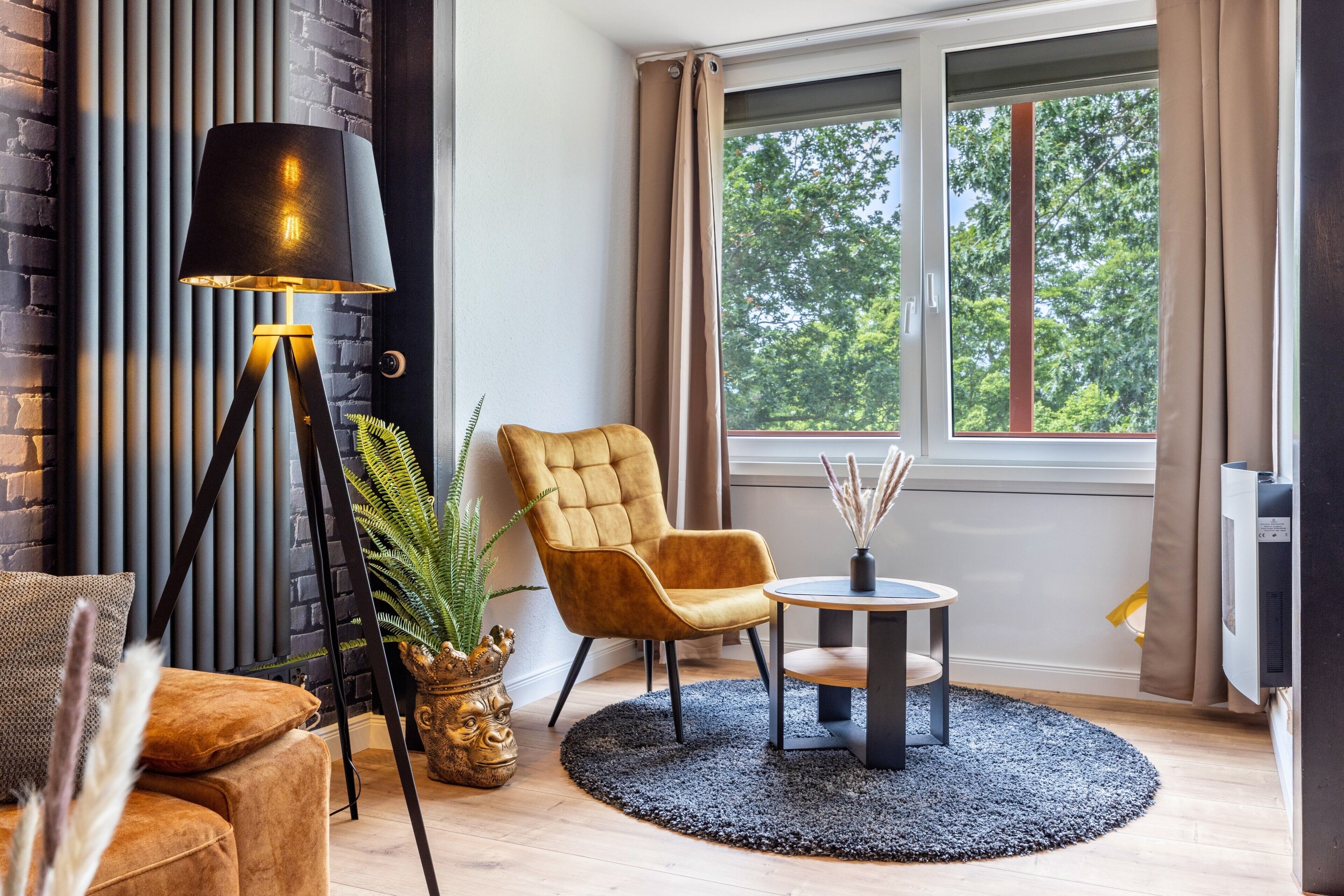 A modern living room with a sofa, armchair, floor lamp, small table, and a large window with curtains.