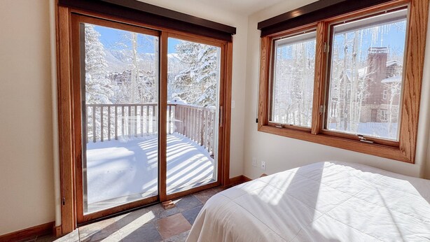 Un dormitorio con una cama grande, un ventanal corredero que da a un balcón con vistas a árboles cubiertos de nieve y un edificio.