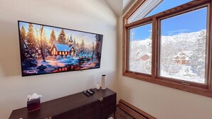 Una habitación con un televisor grande montado en la pared que muestra un paisaje invernal, una ventana de madera con vistas a árboles cubiertos de nieve y un mueble multimedia oscuro con controles remotos y un recipiente de vidrio.
