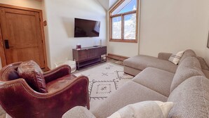 Una sala de estar con un sillón de cuero marrón, un sofá seccional gris, un televisor de pantalla plana montado en la pared y una ventana con vista a árboles cubiertos de nieve.