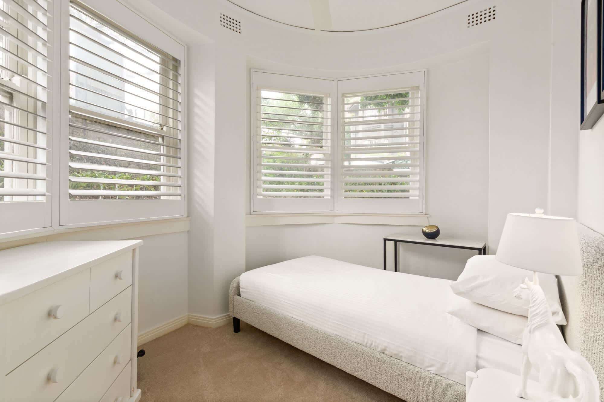Une chambre avec un lit simple, une table de chevet avec une lampe et une commode. Il y a deux fenêtres équipées de stores, dont l’une offre une vue sur la verdure à l’extérieur.