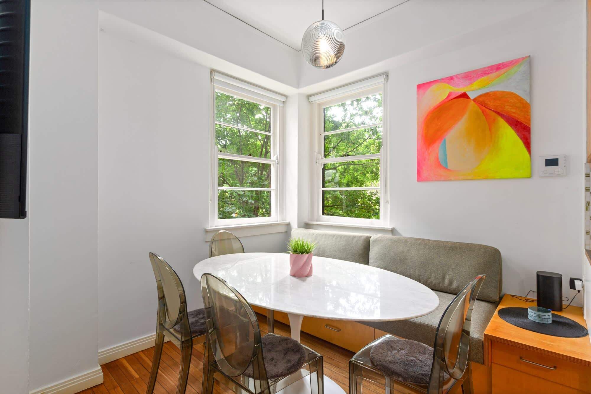 A modern dining room with a round table, clear chairs, a bench, and a large window with a view of trees.
