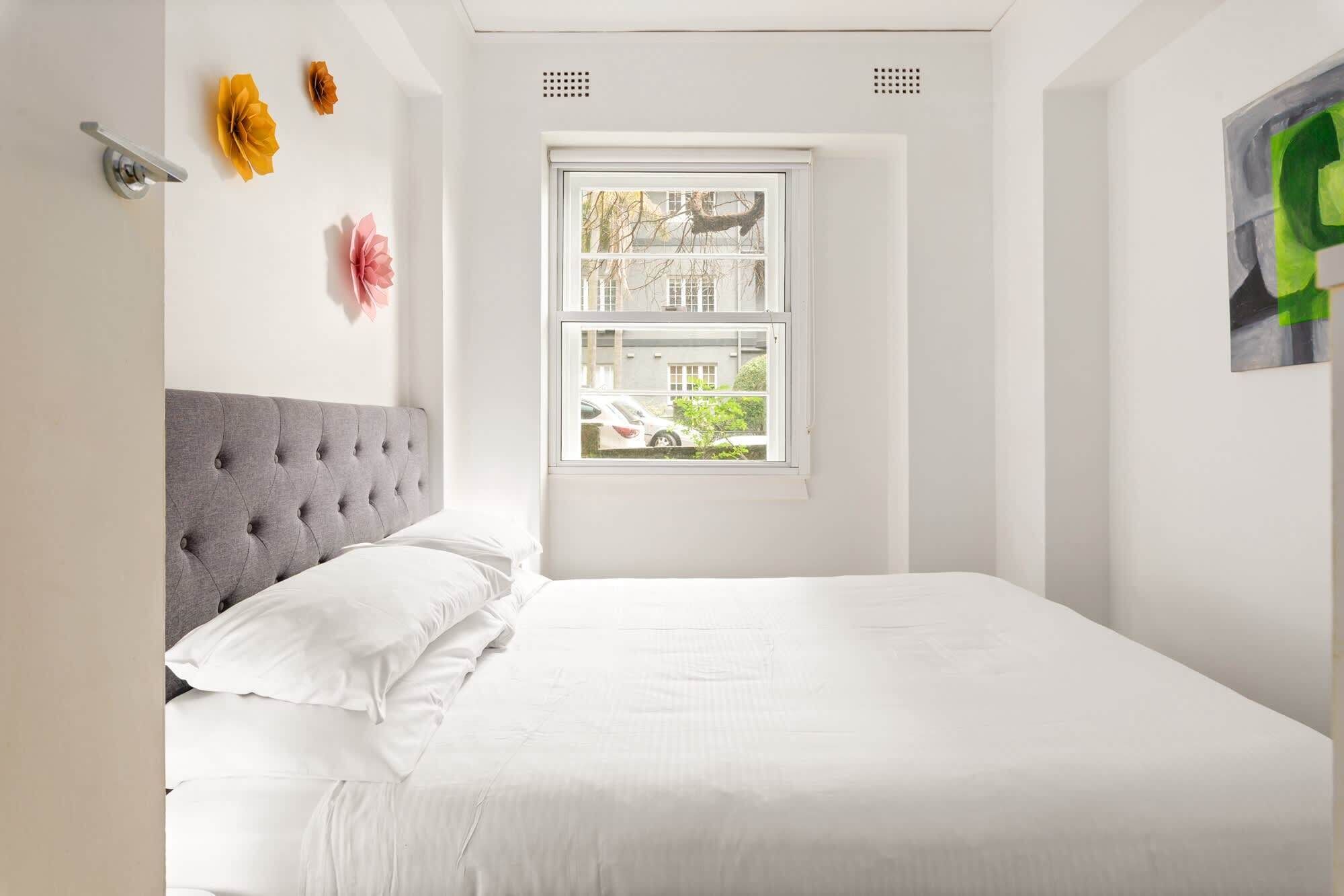 Une chambre à coucher avec une tête de lit capitonnée grise, du linge de lit blanc, une fenêtre donnant sur un immeuble et des voitures garées, et un tableau abstrait vert accroché au mur.