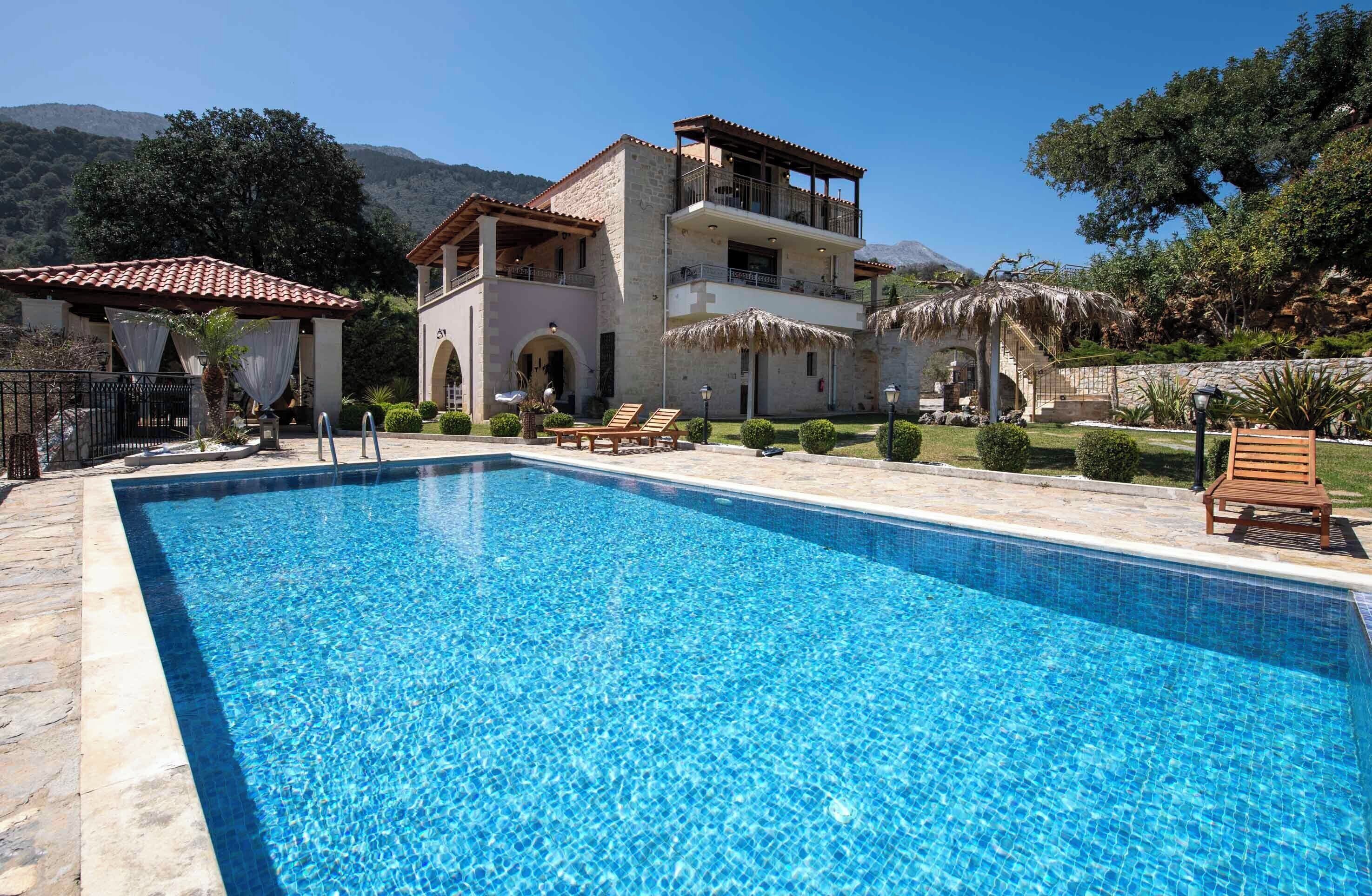 Une grande piscine entourée de chaises longues, un bâtiment à deux étages doté de balcons, et un paysage montagneux en arrière-plan.