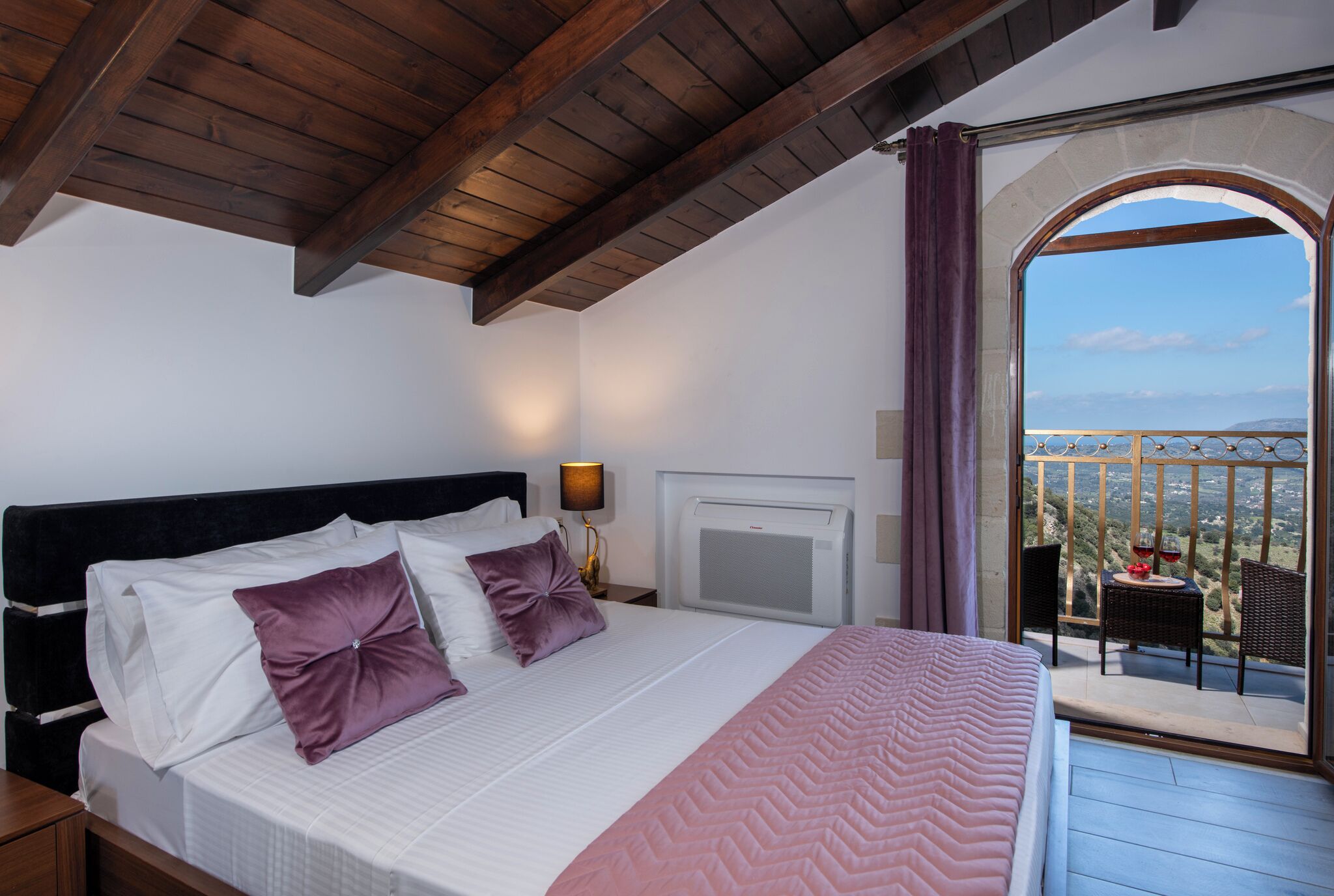 A bedroom with a large bed, a nightstand, a lamp, and a view of the outdoors through an arched window.