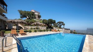A swimming pool with a metal ladder, surrounded by stone tiles. There are wooden lounge chairs and a thatched umbrella. A stone wall and trees are visible in the background.
