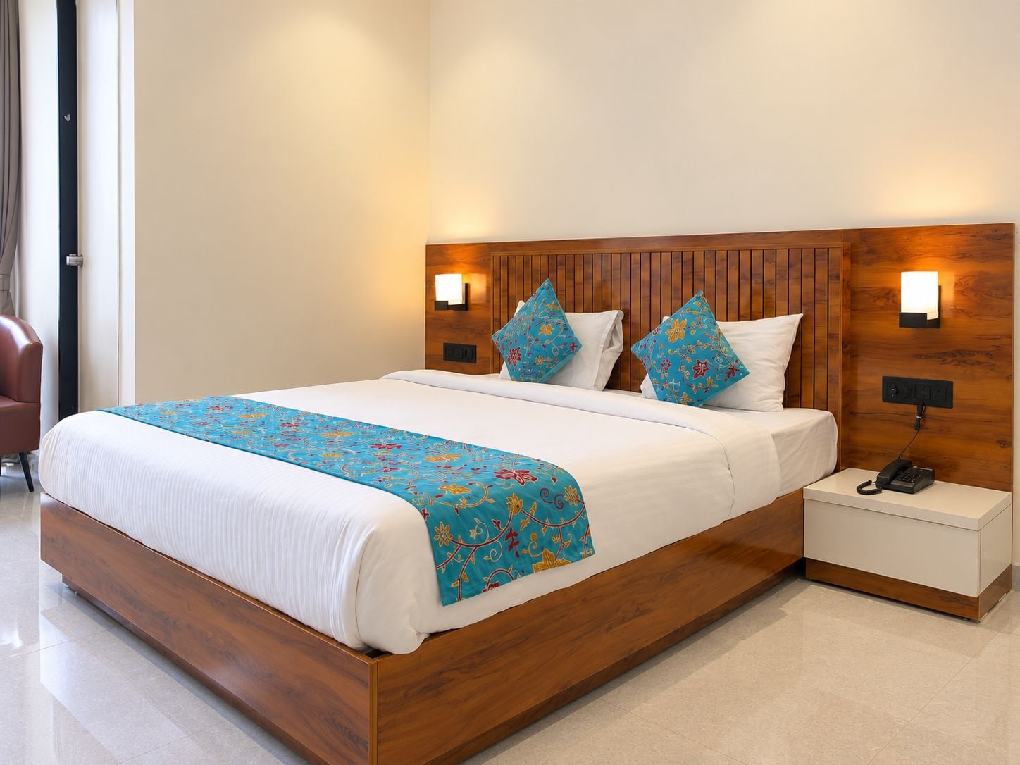 Een hotelkamer met een houten bedframe, een witte dekbedovertrek en twee decoratieve kussens. Er staat een nachtkastje met een telefoon en een wandlamp.