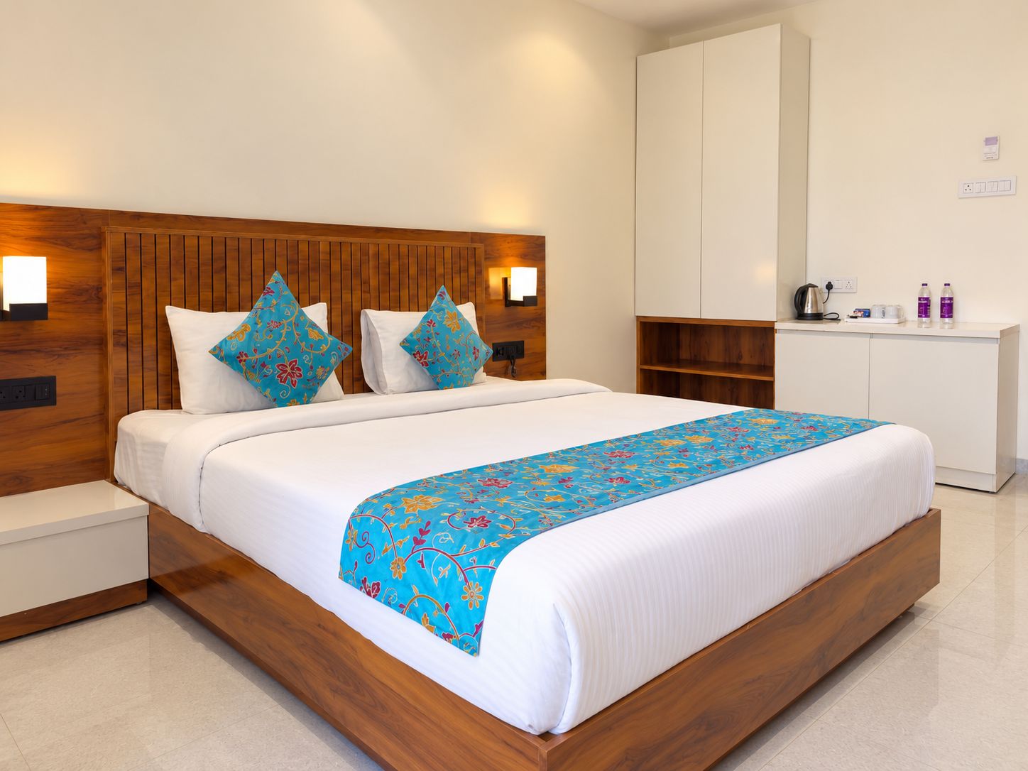 A hotel room with a wooden headboard, a bed with white bedding, a blue floral throw, and a white wardrobe.