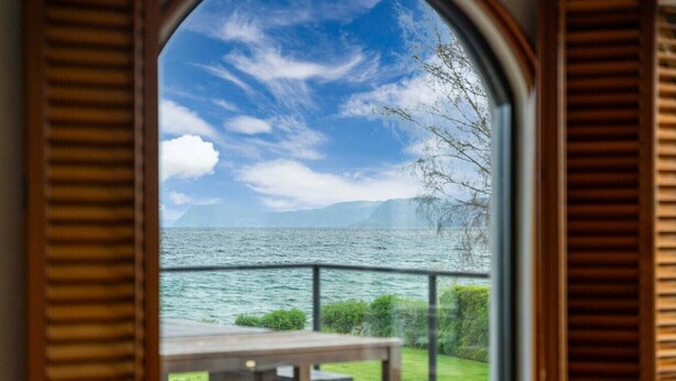 A view from a room with wooden shutters, overlooking a body of water with a grassy area and a wooden table.
