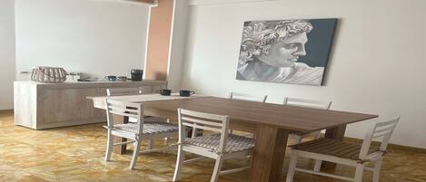 A dining area with a wooden table and white chairs, a painting on the wall, and a kitchen in the background.