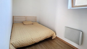 A small, single-bed room with a wooden headboard, a beige patterned bedspread, a single pillow, and a wall-mounted radiator.