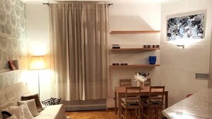 Ett mysigt vardagsrum med en soffa, ett matbord med stolar, ett element och en vägghylla med böcker och en vas.