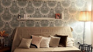 Ett modernt vardagsrum med en mönstrad tapet, en soffa med många kuddar, en hylla med diverse föremål och en lampa.