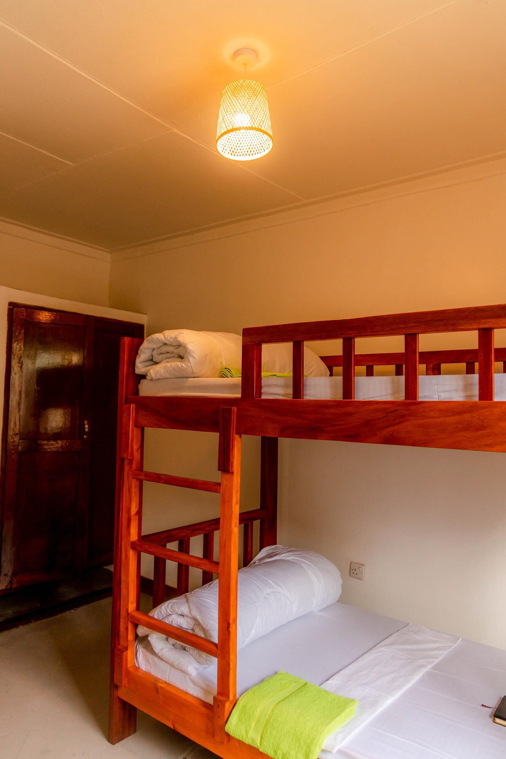 A bunk bed with a wooden frame and white bedding, a single bed with white bedding, a green towel, and a hanging light fixture.