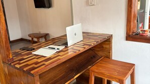 Mesa de madeira com um laptop, um caderno e um smartphone. Há uma televisão fixada na parede e uma janela com vista para o exterior.