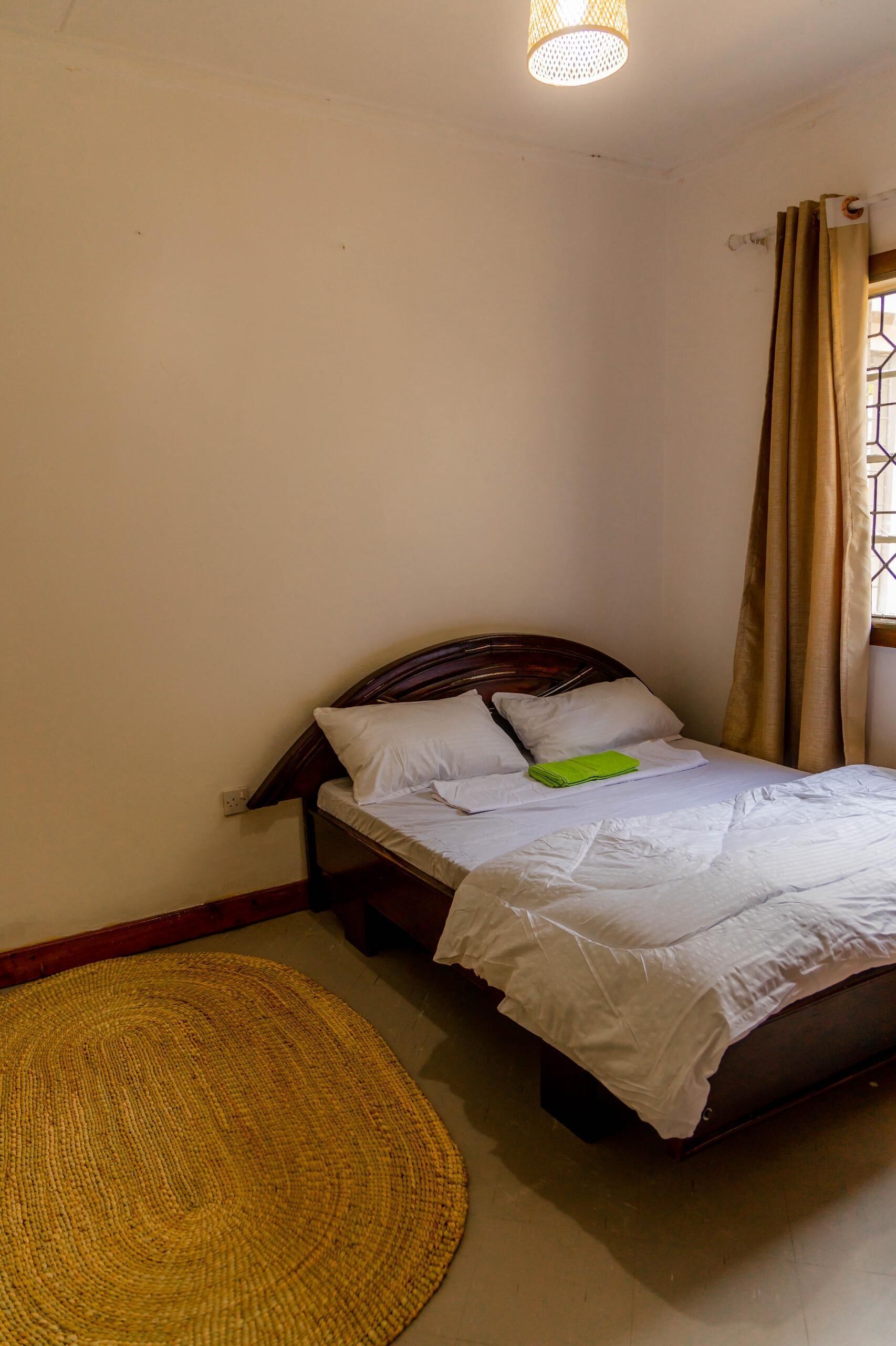 Cama bem arrumada com roupa de cama branca e toalha verde, um tapete tecido e cabeceira de madeira simples.