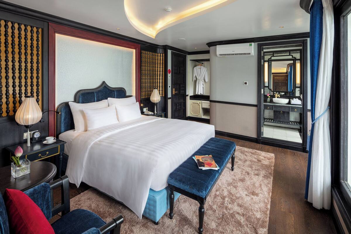 A hotel room with a large bed, bedside tables, a bench, a mirror, and a view of the outside.