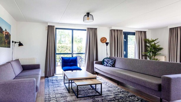 A modern living room with a sofa, a coffee table, a blue chair, and a large window with curtains.