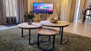 A living room with a TV, two round tables, a bowl of fruit, a plant, and a lamp.