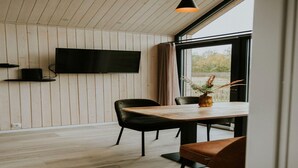 A modern dining area with a wooden table, black and brown chairs, a flat-screen TV mounted on the wall, and a large window with a view of trees. - Charming Tiny House Getaway with Terrace. (Balkbrug)