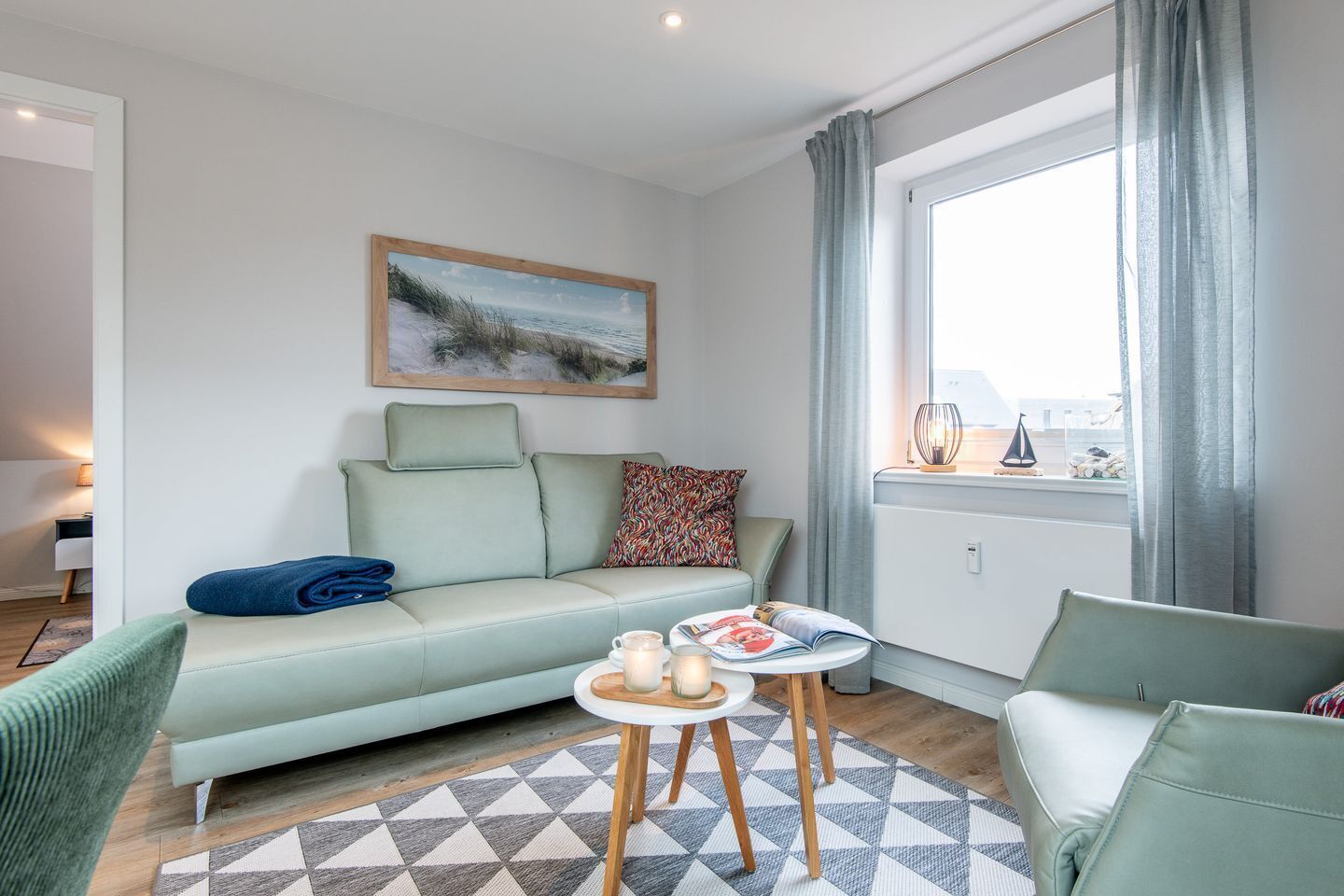 Ein modernes Wohnzimmer mit einem hellgrünen Sofa, einem gemusterten Kissen, einem kleinen runden Tisch mit Kerze und Zeitschrift und Blick auf das Meer durch ein Fenster mit Vorhängen.