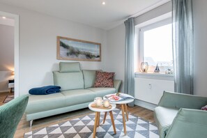 A modern living room with a sofa, a small table with a magazine, a lamp, and a window with curtains.