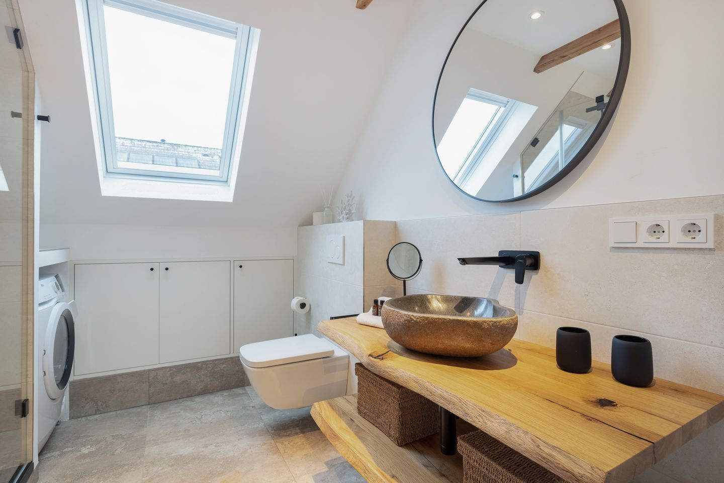 Un baño moderno con tragaluz, espejo redondo, tocador de madera, lavabo de piedra y un inodoro.