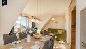 Un comedor con una mesa de madera dispuesta para cuatro personas, rodeada de sillas grises tapizadas. Se ve un sofá verde y una lámpara de pie al fondo.