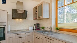 Una cocina moderna con armarios de madera clara, horno empotrado, cafetera y ventana con vistas a árboles.