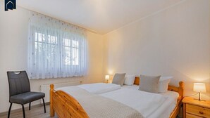 A bedroom with a wooden bed, two bedside tables, a chair, and a window with sheer curtains. - Modern vacation apartment with terrace & garden - relaxation on the Achterwasser - Loddin (Loddin)
