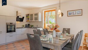 A modern kitchen with white cabinets, a built-in oven, and a dining area with a wooden table and grey chairs. - Modern vacation apartment with terrace & garden - relaxation on the Achterwasser - Loddin (Loddin)
