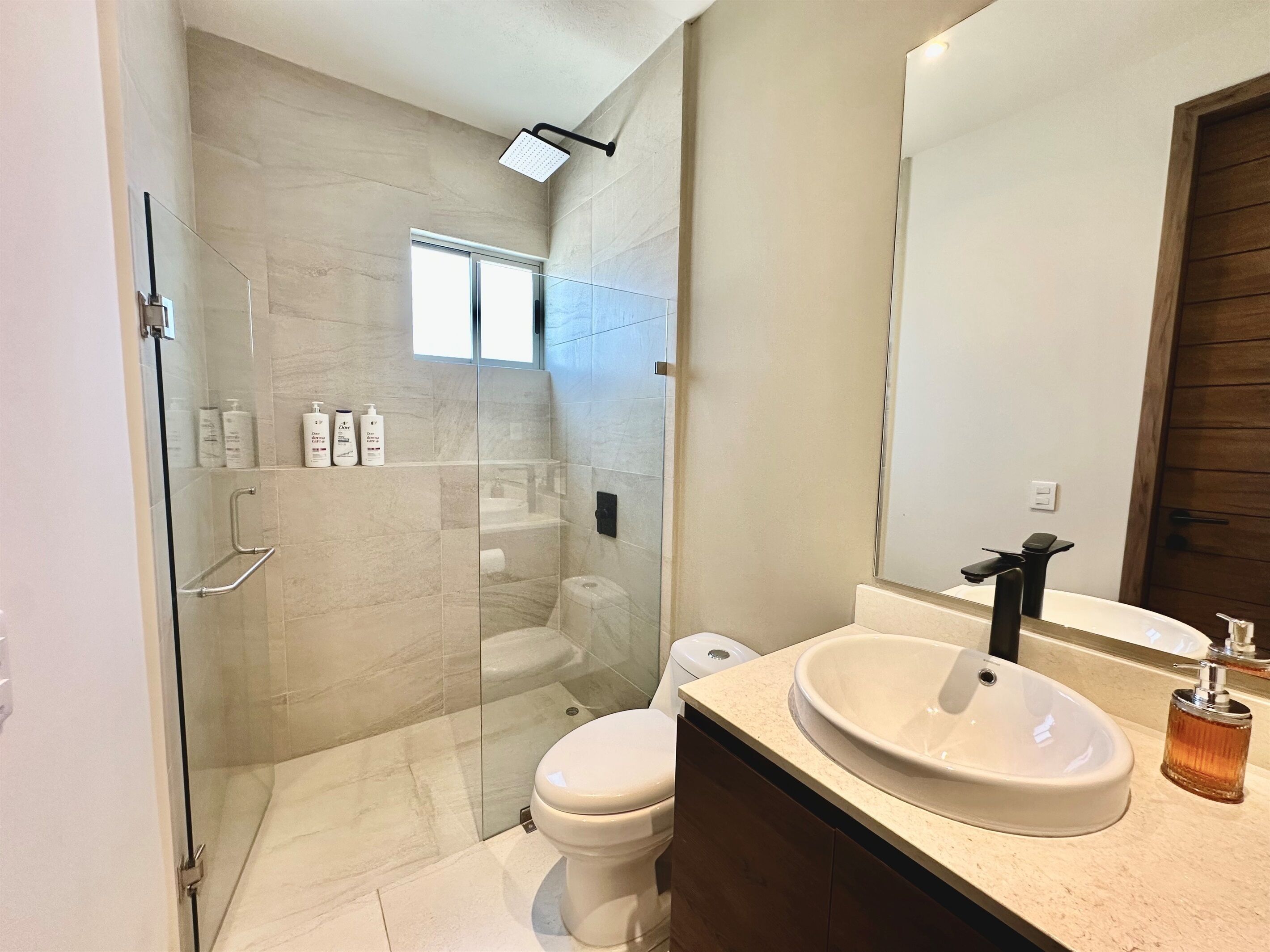Ein modernes Badezimmer mit Duschkabine aus Glas, Toilette, Waschbecken mit Spiegel und Pflegeprodukten an der Wand.