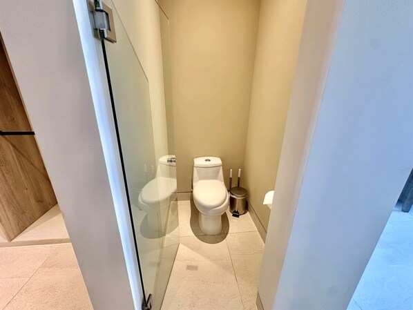 A small, modern bathroom with two toilets, a trash bin, and a glass partition.