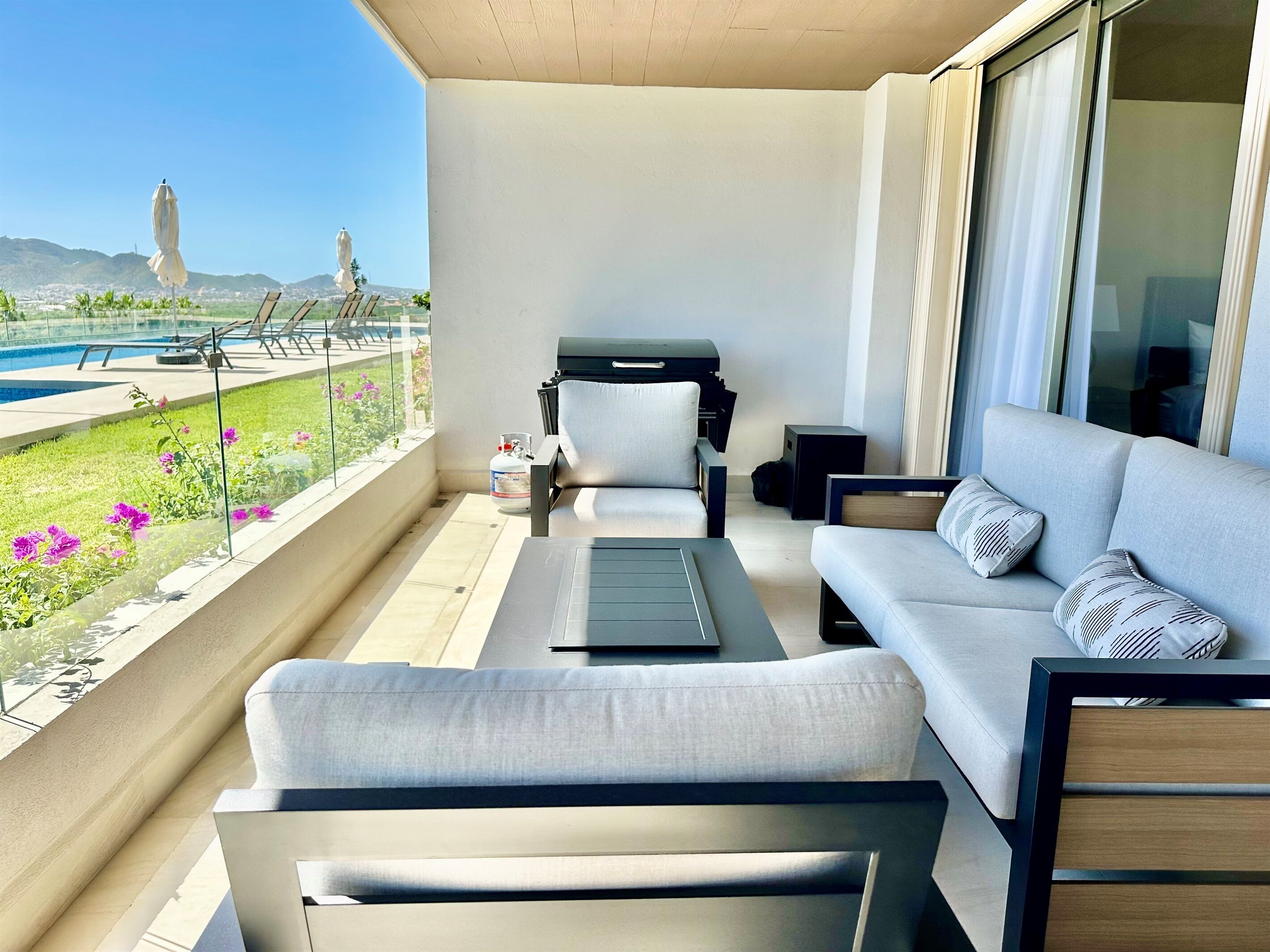 Ein Balkon mit einer Couch, Stühlen, einem Tisch und einem Grill. Im Hintergrund ist ein Pool und Berge zu sehen.