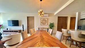 Un'area pranzo moderna con un lungo tavolo in legno, sedie beige, un lampadario e un televisore a schermo piatto montato a parete.