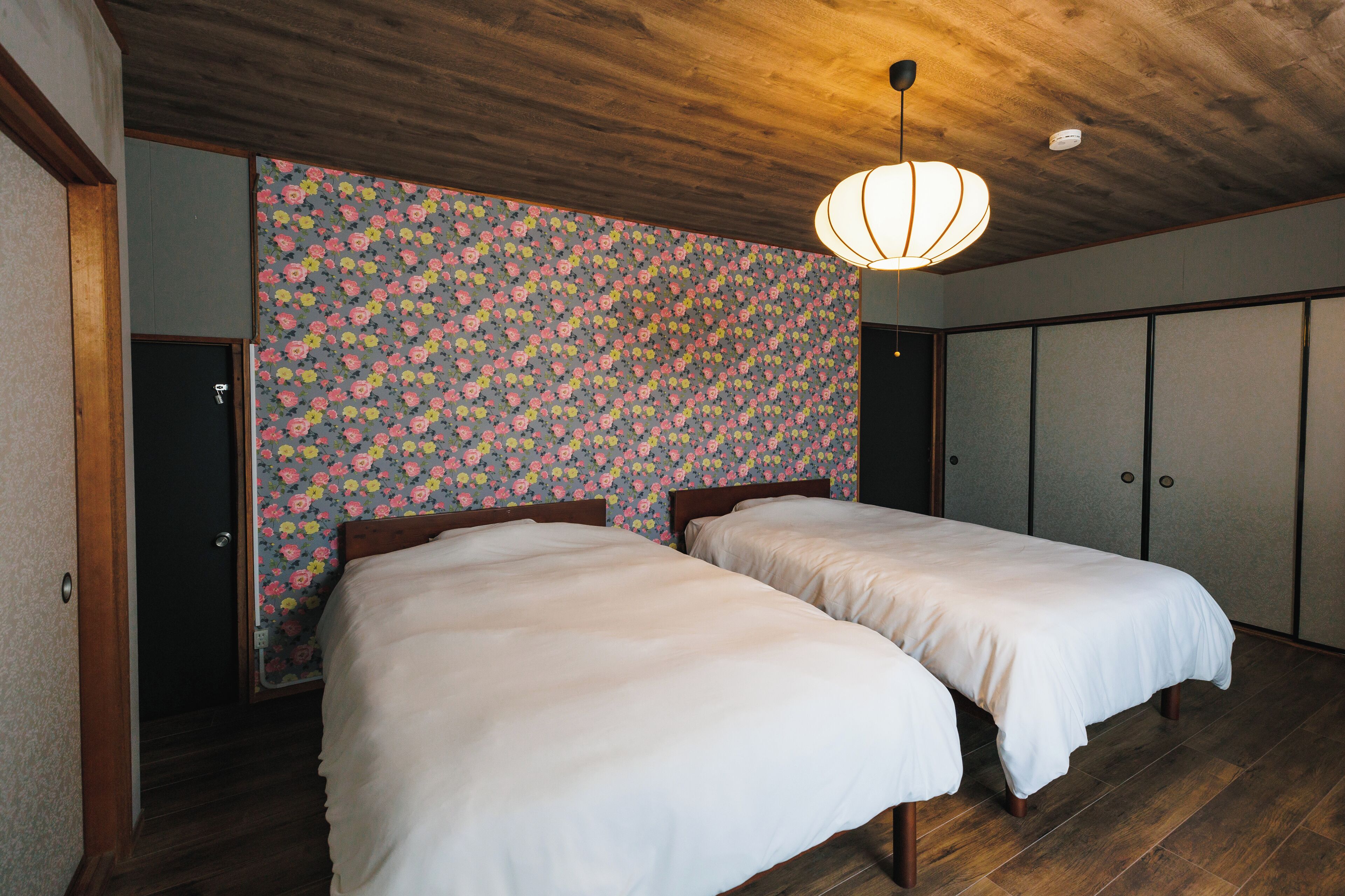 A room with two beds, wooden flooring, a floral wallpaper, and a ceiling light fixture.