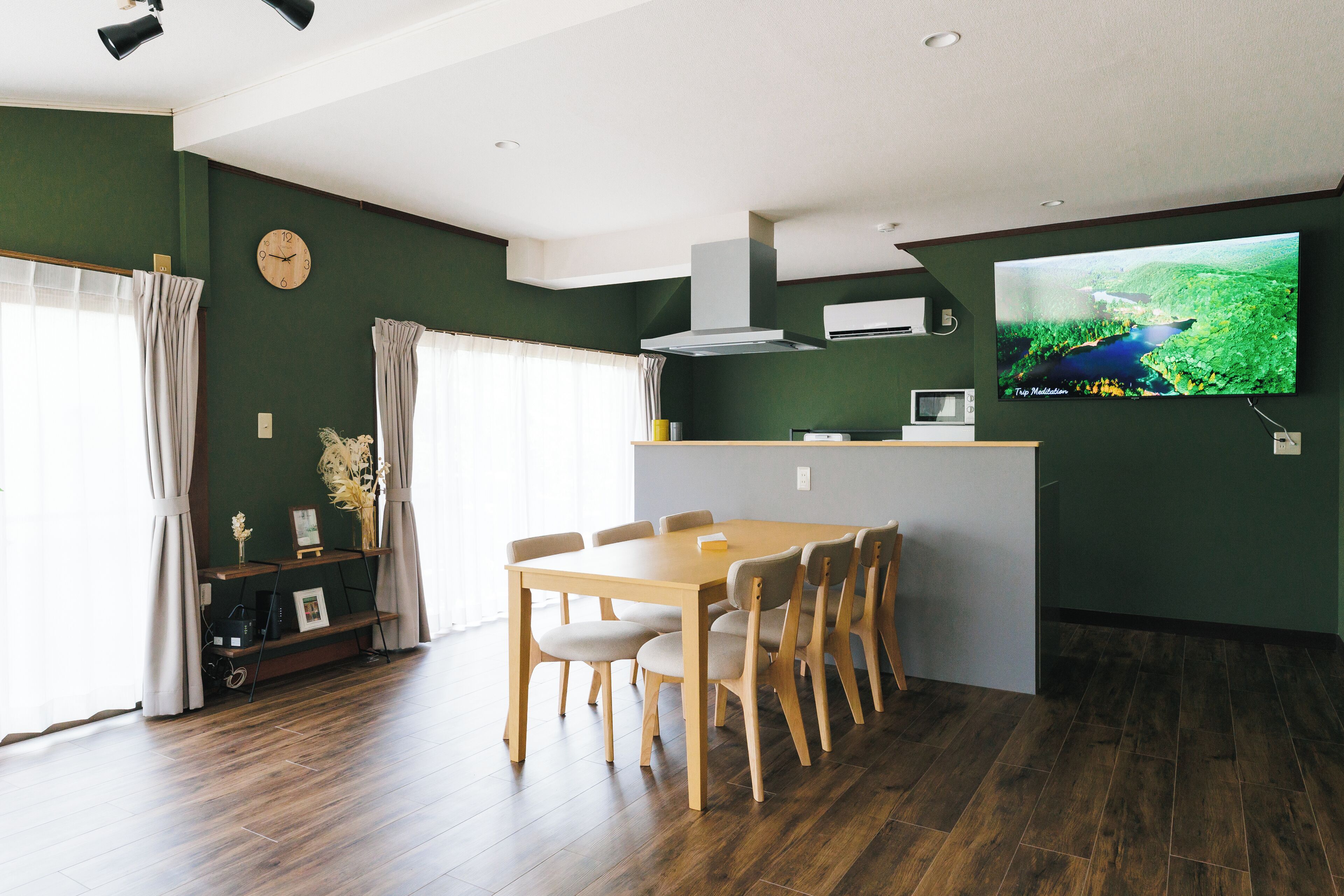 A modern kitchen with a dining area, wooden flooring, and a flat-screen TV on the wall.